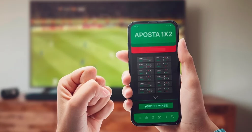 Pessoa segurando smartphone com aposta 1x2 na tela, exibindo opções de apostas esportivas, com TV de futebol ao fundo. Ideal para quem busca dicas de apostas esportivas e aposta 1x2.